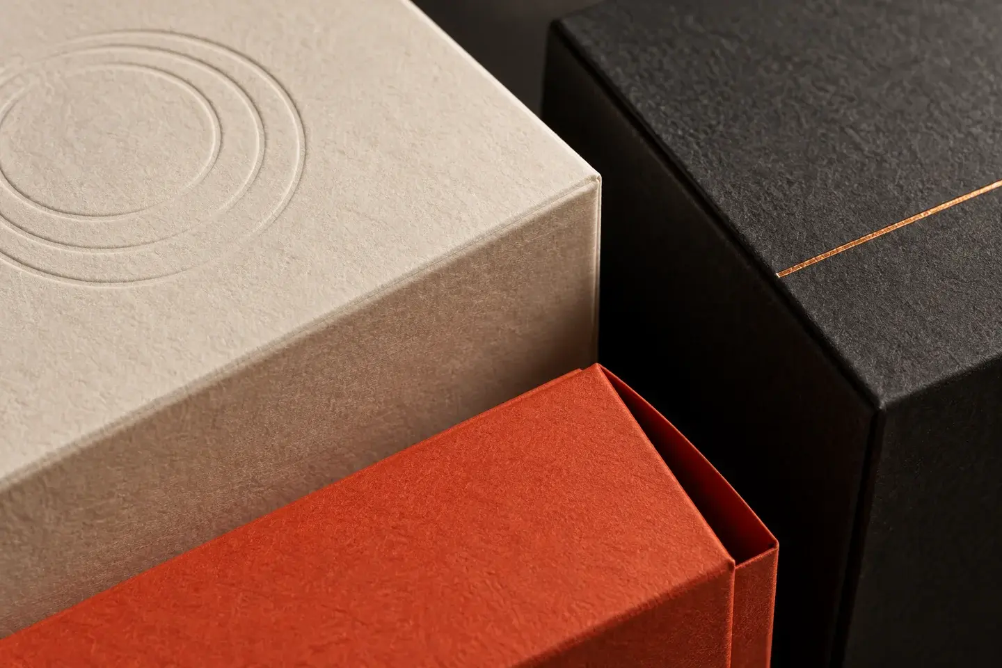 Close-up of ivory textured paper with blind debossing, a fine copper foil line on charcoal paper, and an orange-red sleeve.