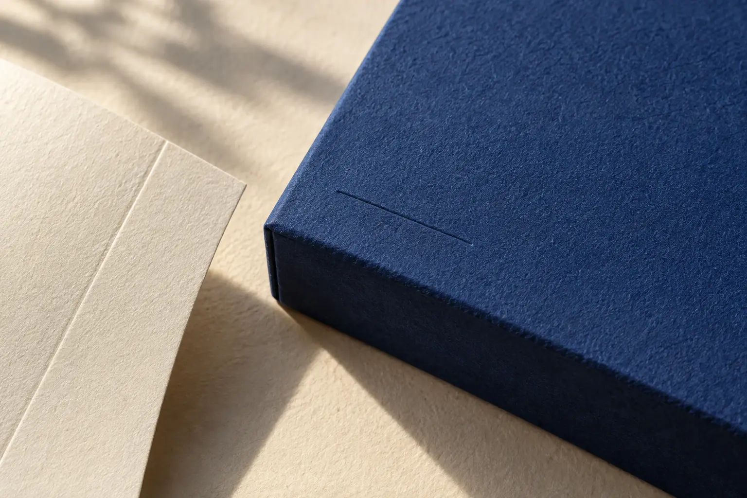 Close view of textured blue paper wrapped around a thick box lid beside an ivory paperboard sample with a crease.
